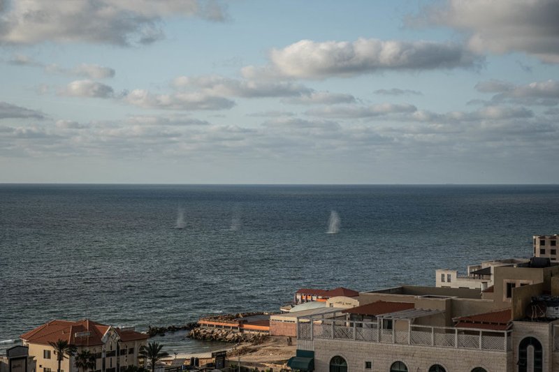 Spray rises from the ocean surface where numerous shells have hit it. In the foreground, pale buildings with brown roofs and palm trees crowd the shoreline. The image is bisected by the horizon.