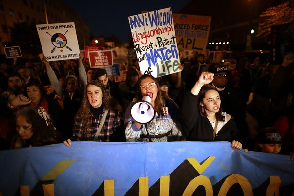 Three women march defiantly holding a blue banner, the middle one is yelling into a speakerphone. It's night time. Signs in the march read 'Respect & Protect Our Water of Life' and 'No Dakota Access Pipeline'