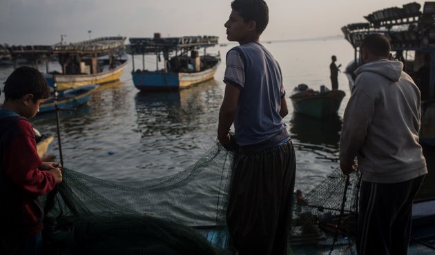 Palestinian fisherfolk, including a small boy, hold a net near the ocean's edge in low light. Several small vessels are visible in the near distance.