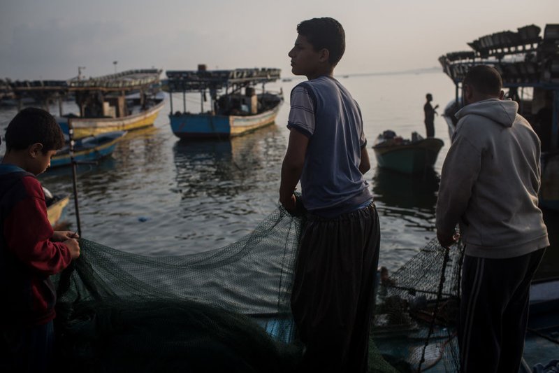Palestinian fisherfolk, including a small boy, hold a net near the ocean's edge in low light. Several small vessels are visible in the near distance.
