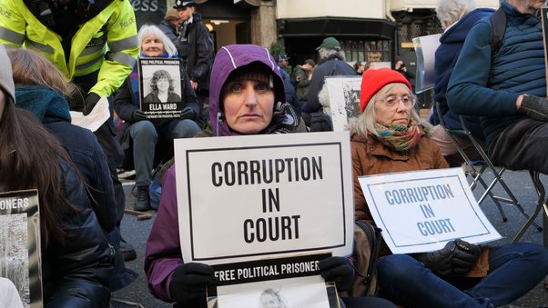 Two women, one middle aged and one elderly, sit sombrely on the pavement with signs reading "corruption in court" in their hands.