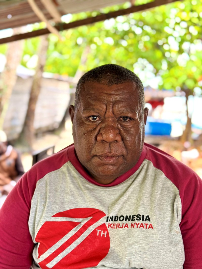 The village chief is wearing a red and white t-shirt and looking steadily into the camera
