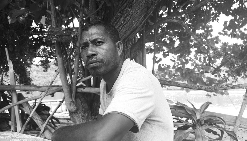 Black and white portrait of Yayah sitting at a table looking into distance, the sea in the background
